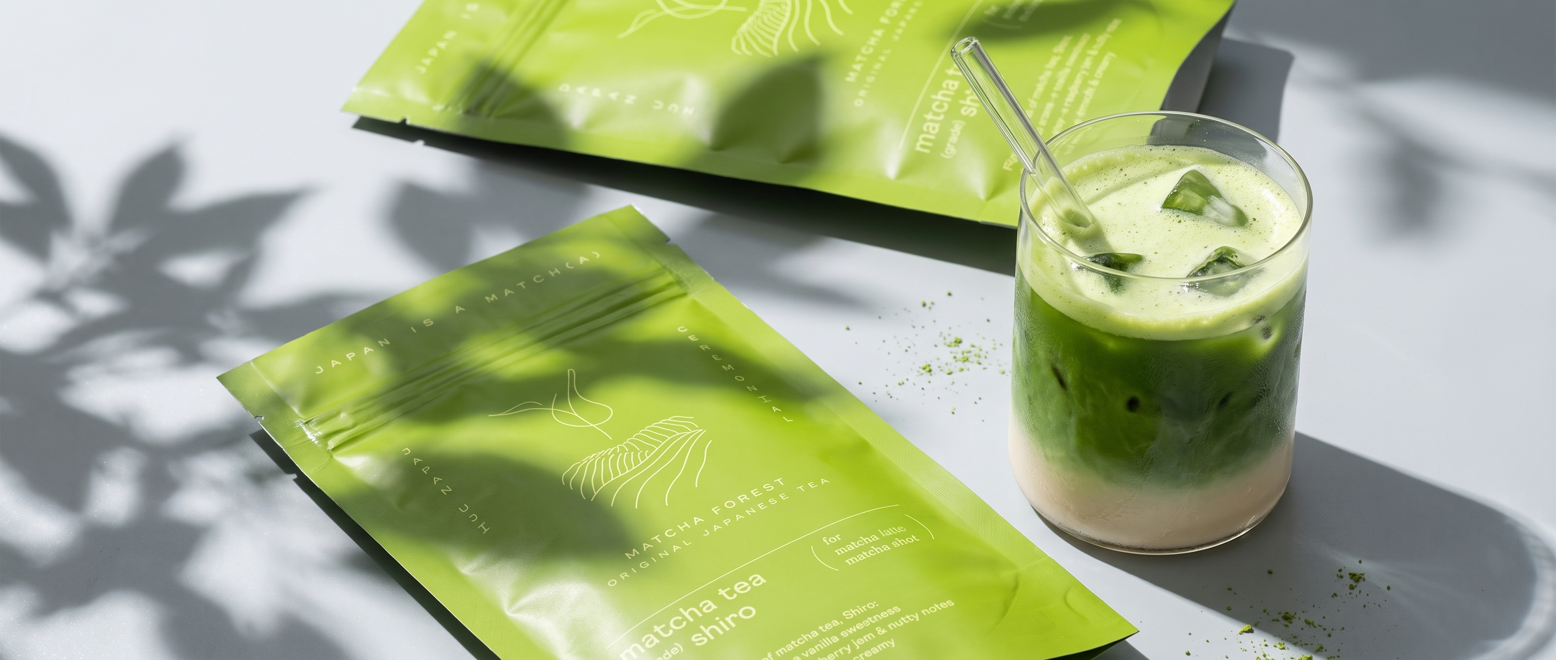 Two bright green matcha forest pouches are arranged on a grey surface with leaf shadows, next to a clear glass of layered green iced matcha latte with a straw.