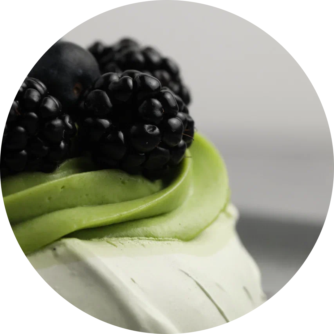 A close-up circular shot of a matcha dessert topped with bright green frosting, blackberries, and a blueberry on a light gray background.