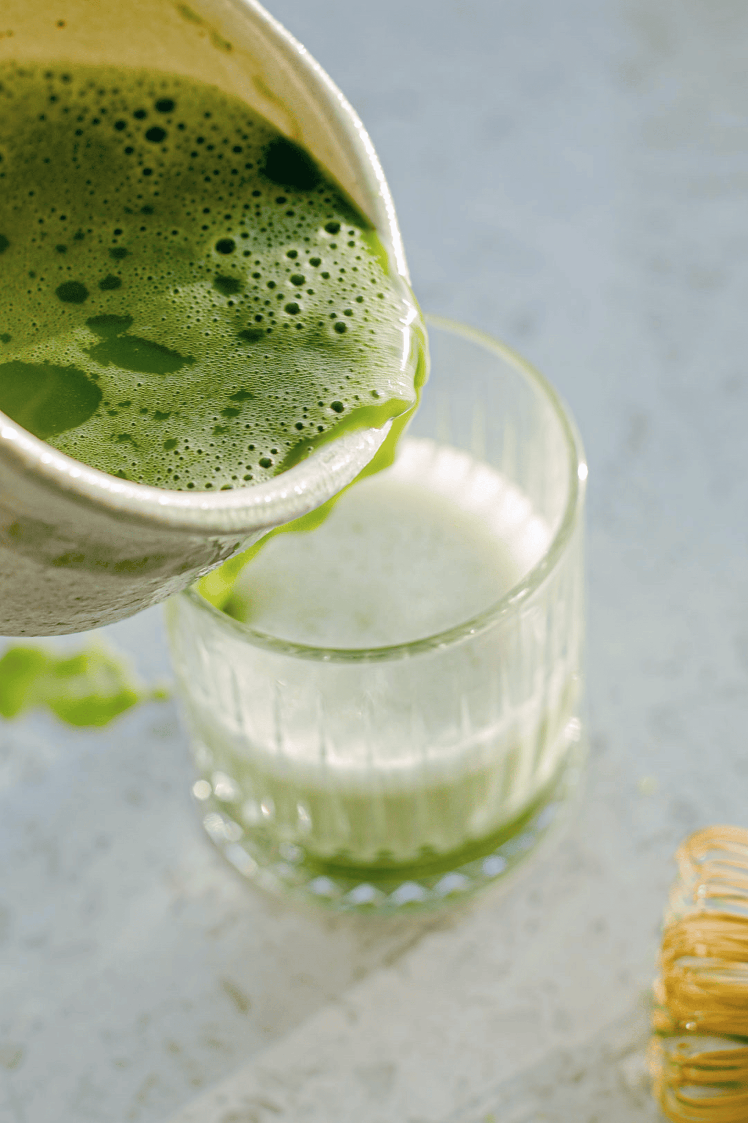 matcha shot being poured from a bowl into a glass of milk on a light grey background