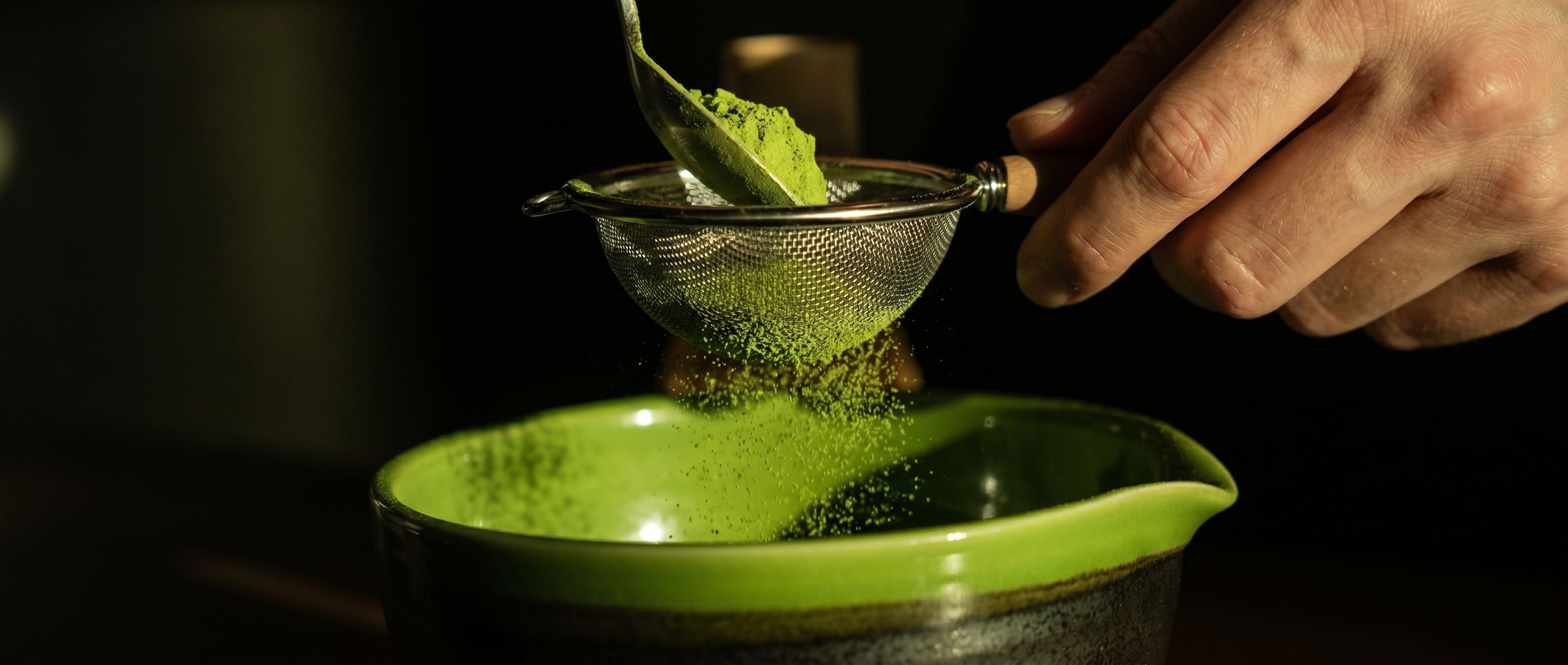 Person sifting matcha green tea powder into a bowl with a dark background