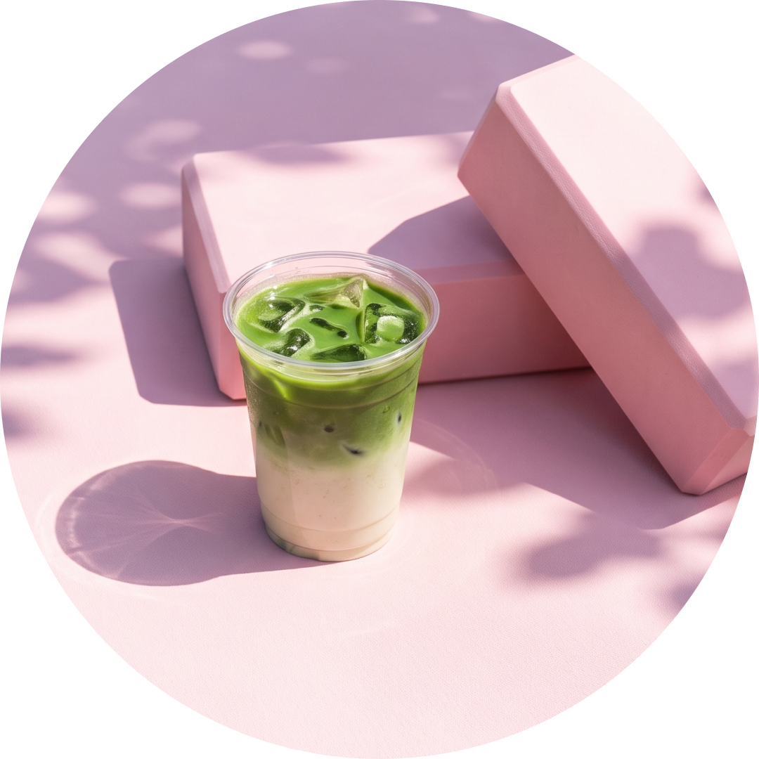 A clear plastic cup filled with an iced matcha latte, placed in front of pink geometric blocks on a pink surface with soft leaf shadows.