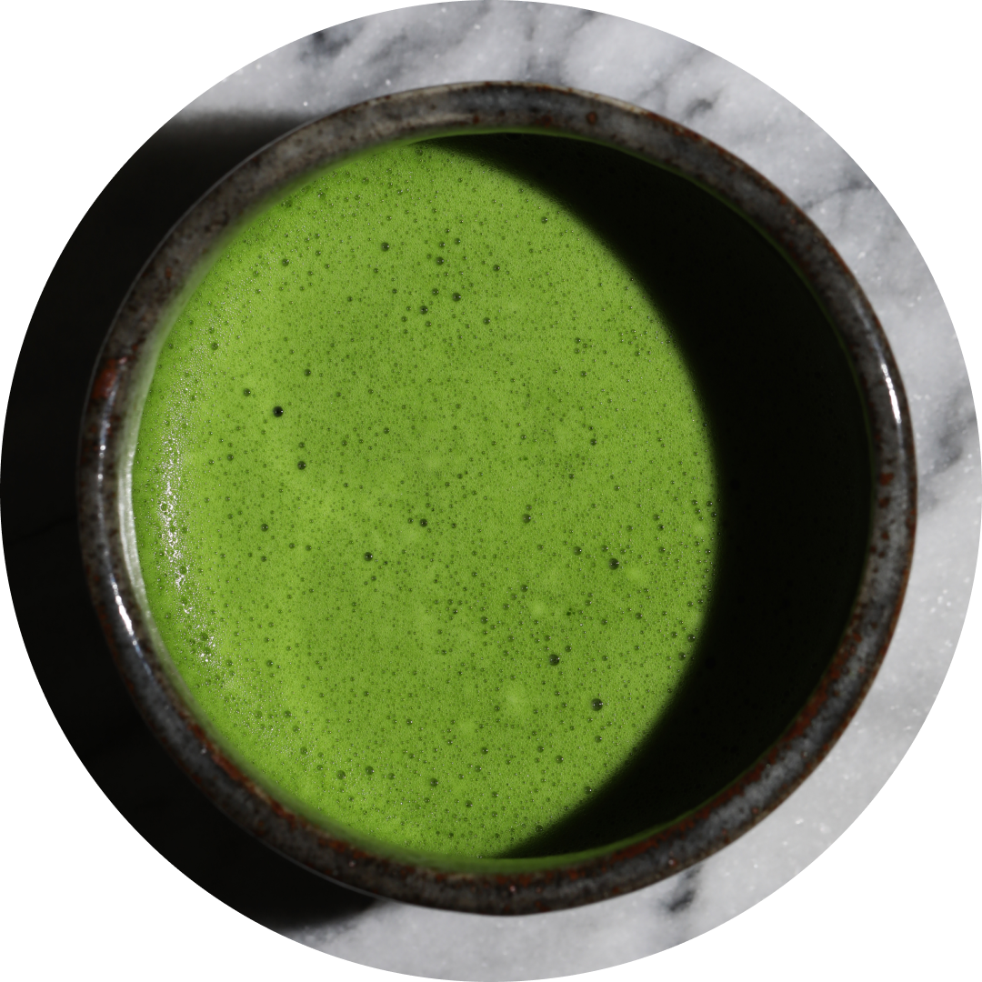 A top-down view of a dark ceramic bowl filled with bright green matcha tea, set on a white marble surface.