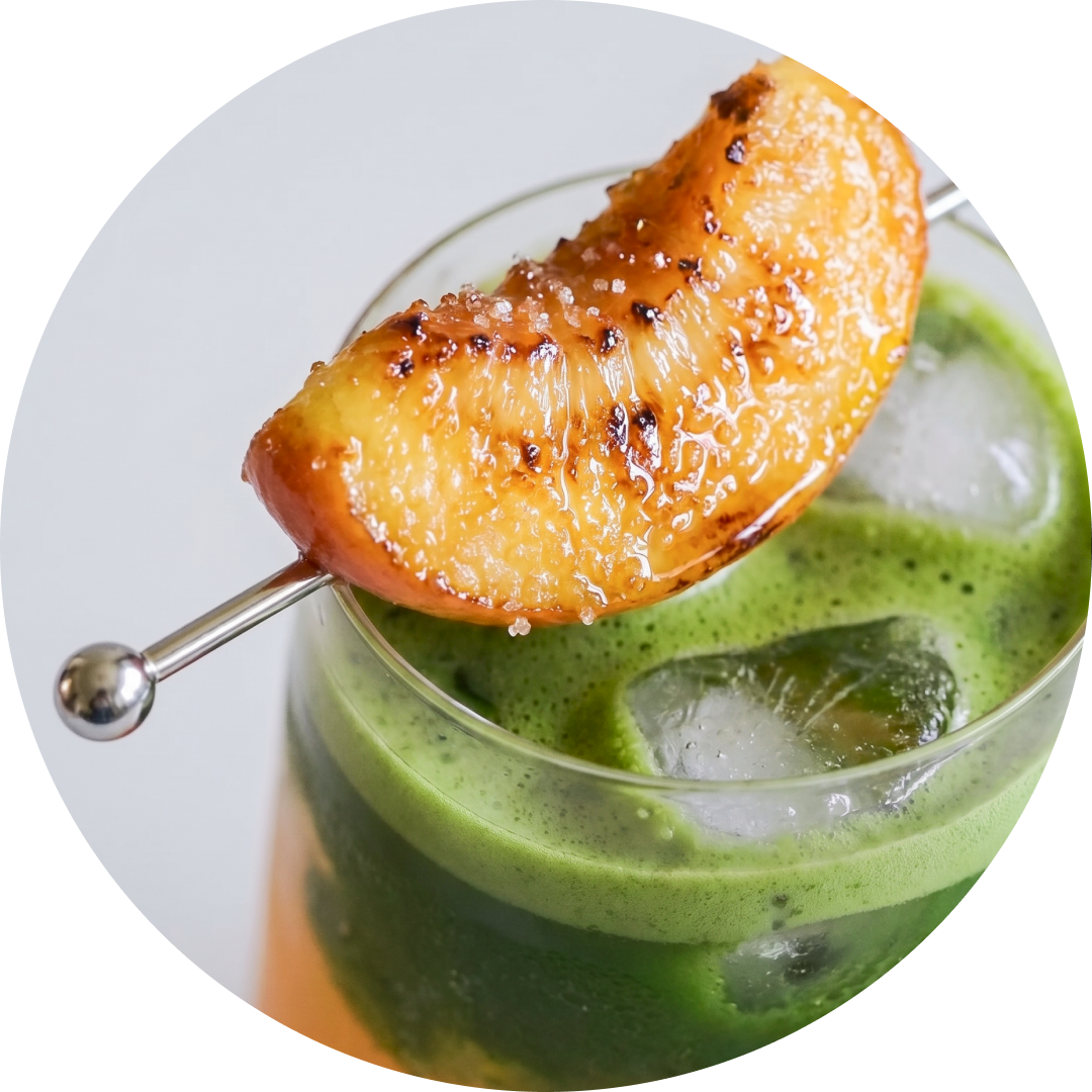 A close-up of a green iced matcha peach latte in a clear glass with ice cubes, topped with a caramelized peach slice on a metal skewer, set against a light gray background inside a circular frame.