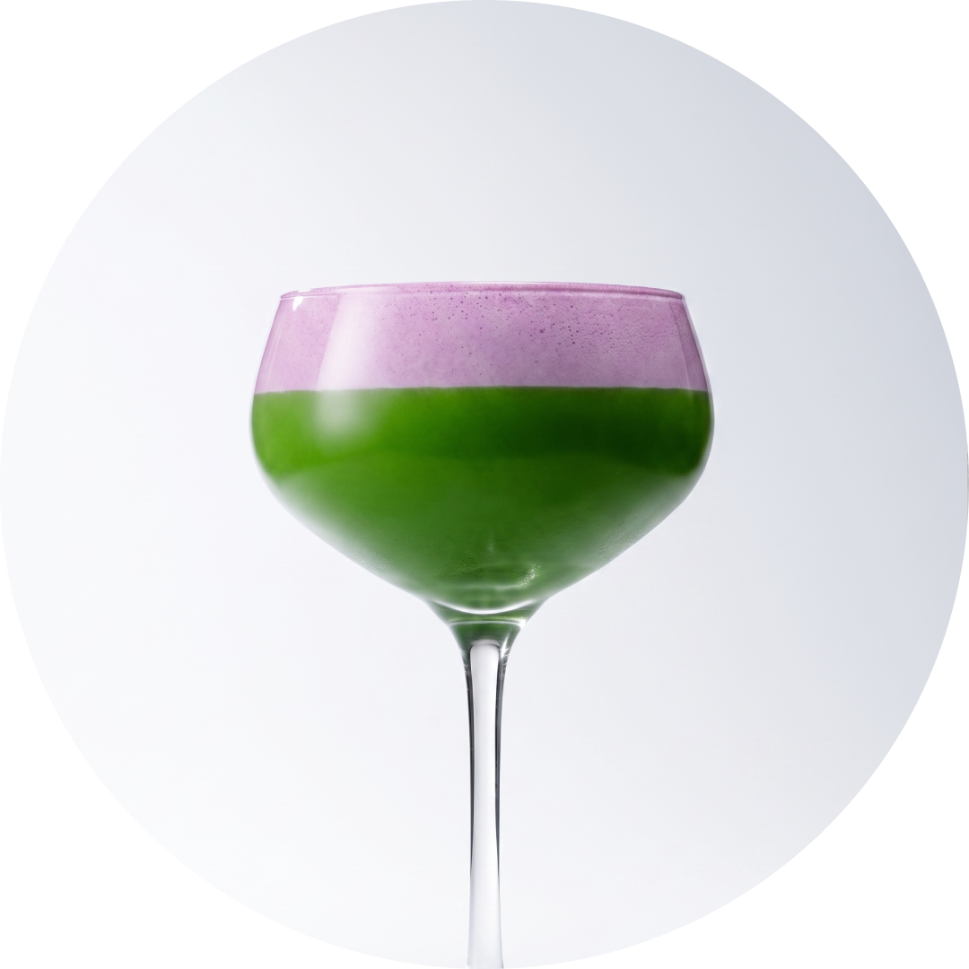 A stemmed coupe glass filled with matcha shot and ube foam, centered against a white circular background.