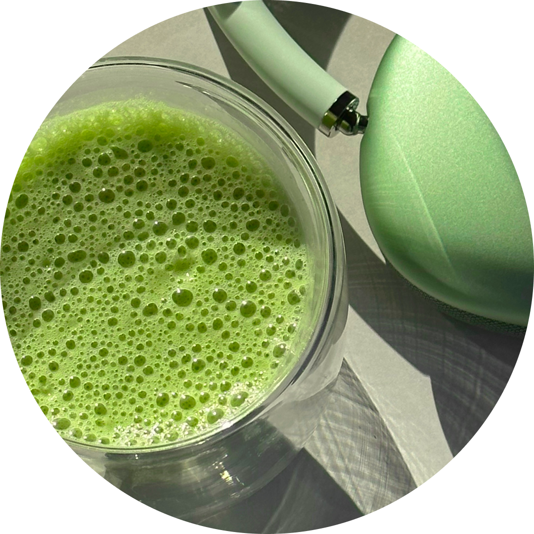 A close-up circular shot of a green matcha smoothie in a clear glass, with part of a mint green headphones on a light surface.